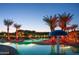 Evening view of a community pool with lit palm trees and a mushroom water feature at 42024 W Rosewood Ln, Maricopa, AZ 85138