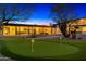 A spacious backyard featuring a putting green, desert landscaping, and an illuminated modern home with a covered patio at 11649 N Miller Rd, Scottsdale, AZ 85260