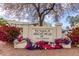 Entrance sign to The Village at Apache Wells 55+ community adorned with colorful flowers and mature landscaping at 5518 E Lindstrom Ln # 3012, Mesa, AZ 85215