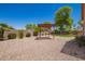 Gravel backyard features desert landscaping, mature tree, plants and a red pergola at 44574 W Garden Ln, Maricopa, AZ 85139