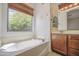 Bright bathroom featuring a soaking tub beneath a window, vanity with dark wood cabinets, and neutral tile flooring at 6941 E Snowdon St, Mesa, AZ 85207