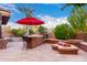 Backyard patio with red umbrella, outdoor kitchen, barstool, firepit bench, and mature desert trees at 17363 W Lisbon Ln, Surprise, AZ 85388