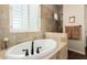 Bathroom featuring tiled walls, travertine flooring, drop in tub with black fixtures and white shuttered window at 3614 W Phillips Rd, San Tan Valley, AZ 85144