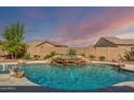 An in-ground pool features a rock waterfall feature, spa, and desert landscaping under a vivid sunset at 18120 W Cassia Way, Goodyear, AZ 85338