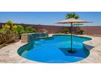 Inviting pool with a waterfall feature, surrounded by a decorative stone patio, lush landscaping, and a shade umbrella at 25546 N 164Th Dr, Surprise, AZ 85387