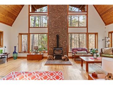 Bright living room with a brick fireplace, wood floors, and soaring windows showcasing the wooded surroundings at 735 Jordan Hills Loop, Chapel Hill, NC 27517