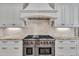 Close up of a gas cooktop and oven with a custom white range hood and stone backsplash at 628 Carolina Crossings Dr, Apex, NC 27523