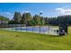 A fenced blue tennis court sits ready for play, with ample lighting and verdant green surroundings under a clear sky at 67 Crimson Way, Pittsboro, NC 27312