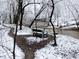 Winter scene of a park bench near a creek, framed by snow-covered trees and a winding trail at 12088 Burlington Rd, Hurdle Mills, NC 27541