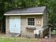 Small wooden outbuilding, painted beige, with a white door, window, and a 