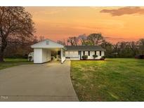 Charming single-story home featuring a connected carport, manicured lawn, and a welcoming front porch under an orange sunset sky at 118 Tysor Cir, Graham, NC 27253