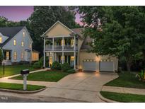 Charming yellow two-story home with double front porch and well-manicured lawn and a two-car garage at twilight at 10415 Ashmead Ln, Raleigh, NC 27614