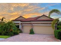 Charming home featuring a red tile roof, stone accents, lush landscaping, and a paver driveway, all under a vibrant sunset sky at 20909 Loggia Ct, Venice, FL 34293