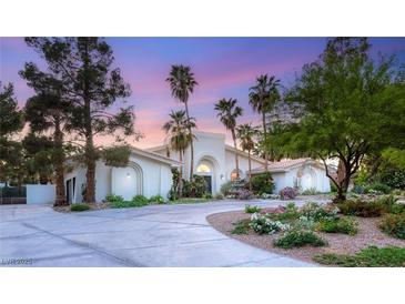 Elegant white home with arched entryway, lush landscaping, palm trees, and a circular driveway at sunset at 7185 Ranchito Cir, Las Vegas, NV 89120