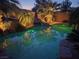 Relaxing pool area featuring a rock waterfall, lush plants, and ambient lighting for evening enjoyment at 10519 Bella Camrosa Dr, Las Vegas, NV 89141