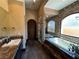 Elegant bathroom featuring a soaking tub with travertine tile surround, arched window, and marble countertop sink at 1800 Amarone Way, Henderson, NV 89012