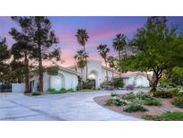 Elegant white home with arched entryway, lush landscaping, palm trees, and a circular driveway at sunset at 7185 Ranchito Cir, Las Vegas, NV 89120